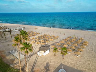 Strand mit Sand, Palmen, Sonnenschirmen und ruhigem blauem Meer unter leicht bewölktem Himmel.