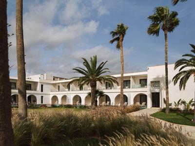 Edificio alberghiero bianco con archi, circondato da palme e prato sotto un cielo azzurro