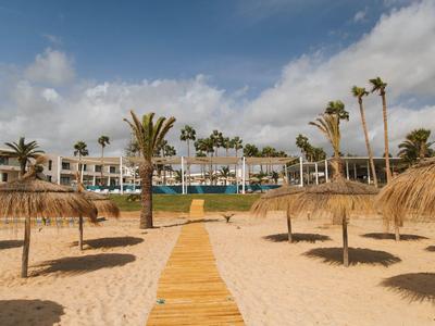 Strand mit Sand, Strohdächern und Holzweg vor einem Hotel mit Palmen unter bewölktem Himmel.