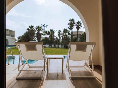 Dos tumbonas y una mesa pequeña en un porche con vista a una piscina y palmeras bajo un cielo azul.