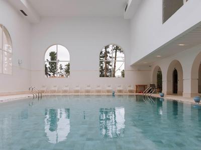 Innenliegendes Schwimmbad mit klarem Wasser, weißen Wänden und großen Rundbogenfenstern.