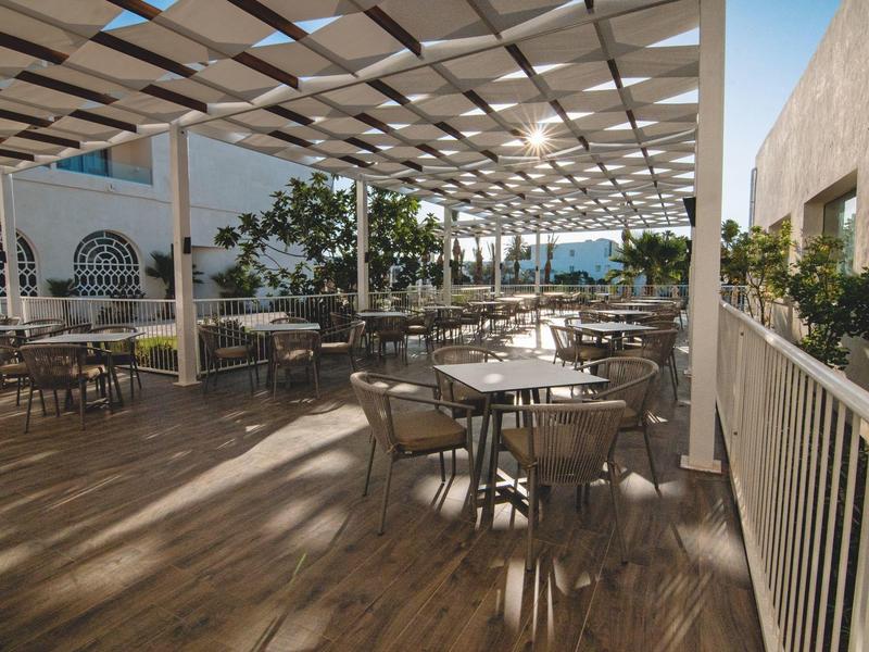 Terraza con mesas y sillas bajo un techo de celosía durante el día en un hotel.