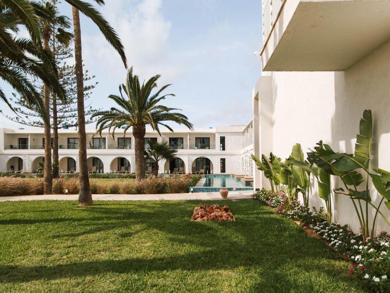Jardín del hotel con palmeras, césped verde y edificio blanco bajo un cielo soleado.