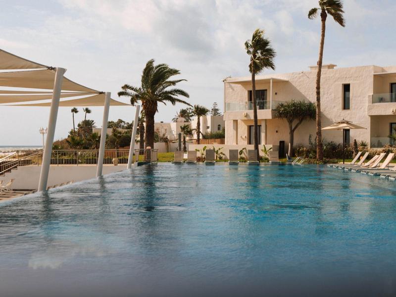 Piscina moderna con tumbonas y palmeras junto a un hotel bajo un cielo nublado.