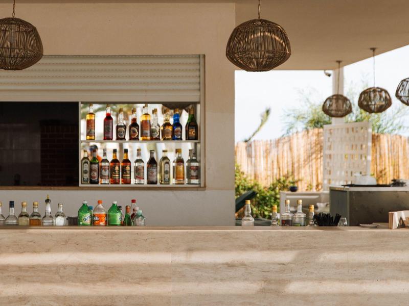 Área moderna de bar de hotel con taburetes, estante de botellas y lámparas colgantes de mimbre.