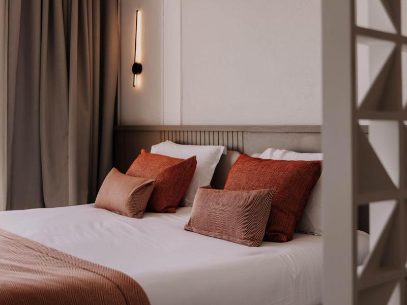 Habitación de hotel moderna con cortinas grises, cojines marrones y cama blanca sencilla.