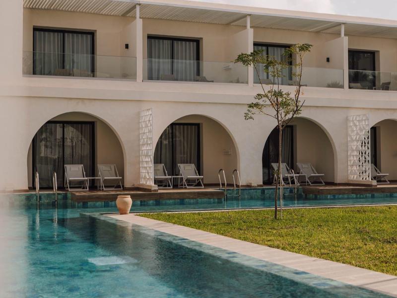 Piscina moderna del hotel con tumbonas frente a habitaciones con grandes ventanas de arco y área verde.
