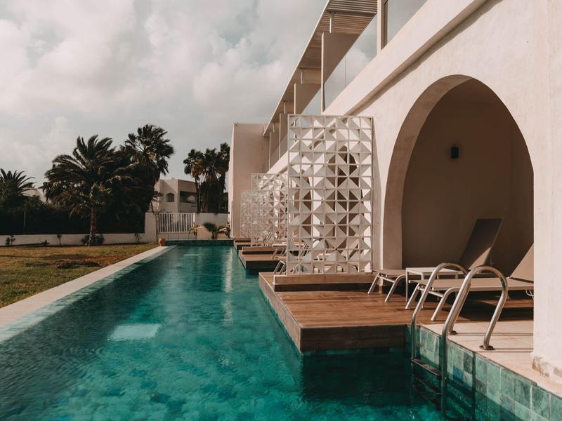 Piscina moderna junto a un hotel con arcos blancos y tumbonas bajo cielo nublado.