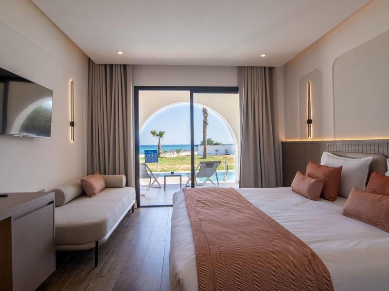 Habitación de hotel moderna con cama doble, sofá y terraza con vista al mar a la luz del día.