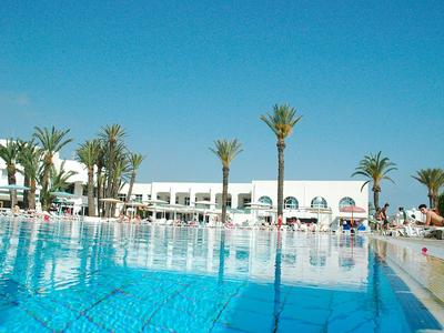 Großer blauer Pool vor weißem Hotelgebäude mit Palmen und klarem Himmel.