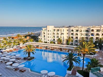 Hotel mit großem Pool, Palmen und weißem Gebäude am Meer unter blauem Himmel.