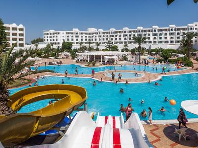 Großer Poolbereich mit Wasserrutschen, vielen Gästen und dahinter einem weißen Hotel bei klarem Himmel.