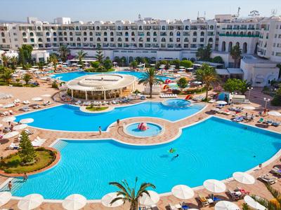 Großer Hotelpool mit Inselbar, umgeben von Liegestühlen und einem weißen Gebäudekomplex.