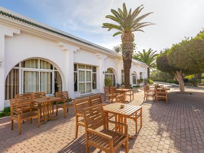 Außenbereich eines Hotels mit weißer Fassade, Holzmöbeln und Palmen auf gepflastertem Boden.