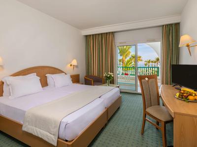 Helles Hotelzimmer mit Doppelbett, Schreibtisch, TV und Balkon mit Blick auf Palmen.