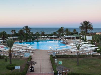 Blick auf einen Pool mit Liegestühlen und Palmen am Meer bei Sonnenuntergang in einem Hotel.