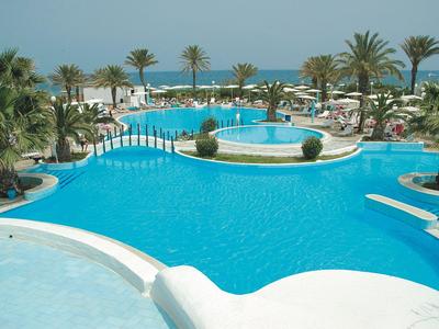 Großer Hotelpool mit Palmen und Meer im Hintergrund unter klarem Himmel.