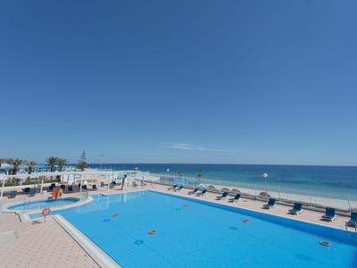 Großer Pool mit Liegestühlen neben dem Strand und klarem blauen Himmel am Urlaubsort.