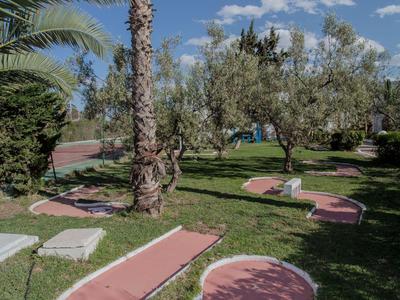Ein sonniger Garten mit Palmen und grünen Bäumen neben einem pinken Minigolf-Pfad unter blauem Himmel.
