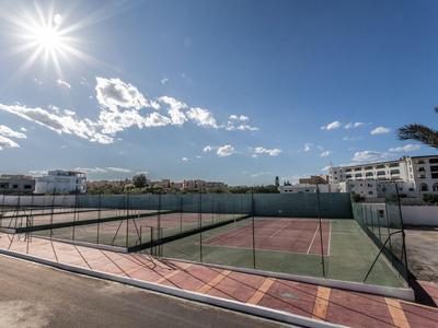 Sonniger Tennisplatz mit Umzäunung und Hotelgebäuden im Hintergrund unter blauem Himmel.