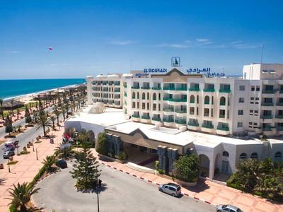 Ein großes Hotelgebäude an der Küste mit Blick auf das Meer und eine palmengesäumte Promenade.
