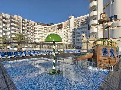 Großer Pool mit Piratenschiff-Wasserspiel und Liegestühlen vor weißem Hotel bei klarem Himmel.