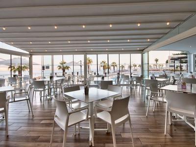 Helles, modernes Restaurant mit Glasfront, weißen Stühlen, Holzboden und Meerblick bei Sonnenuntergang.