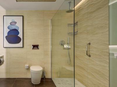 Moderner Duschbereich mit Glaswand, Regendusche, beige Fliesen und Toilette neben einem gerahmten Bild.