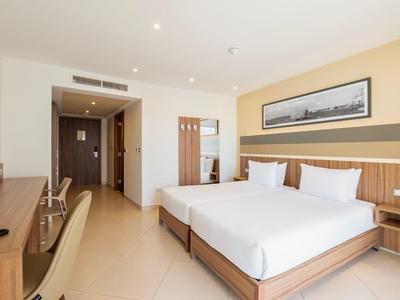 Helles Hotelzimmer mit zwei Einzelbetten, Holzmöbeln und Deckenbeleuchtung.