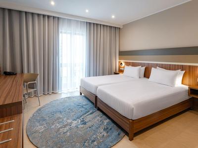 Modernes Hotelzimmer mit zwei Einzelbetten, grauem Teppich, Holzmöbeln und großem Fenster mit Vorhängen.