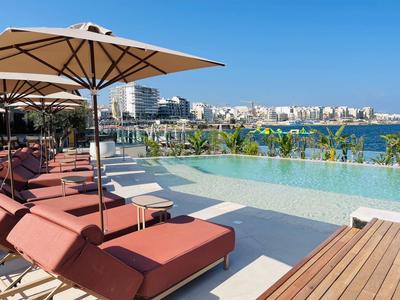 Poollandschaft mit roten Liegen, Sonnenschirmen und Blick auf Meer und moderne Gebäude bei klarem Himmel.