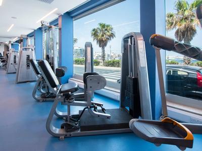 Helles Fitnessstudio mit modernen Trainingsgeräten und großen Fenstern, die Palmen und Auto zeigen.