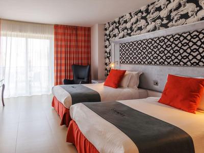 Hell eingerichtetes Hotelzimmer mit zwei Einzelbetten, roten Akzenten und großem Fenster mit Gardinen.