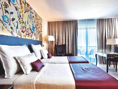 Modernes Hotelzimmer mit Doppelbett, buntem Wandmuster, Vorhängen und Schreibtisch am Fenster.