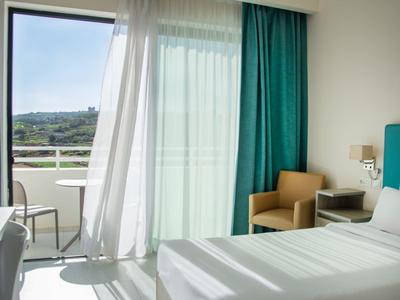 Helles Schlafzimmer mit doppeltürigem Balkon, weißen Wänden, teal-vorhängen und brauem Sessel.