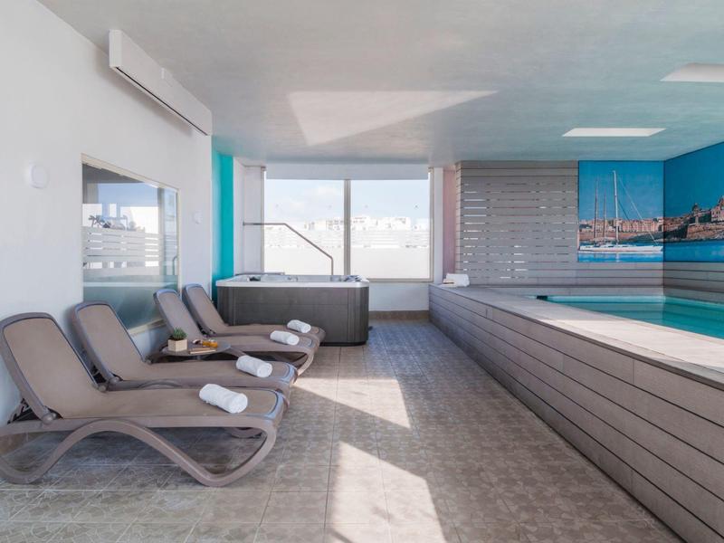 Innenraum eines modernen Hallenbads mit blauen Wänden, Liegestühlen und großen Fenstern.