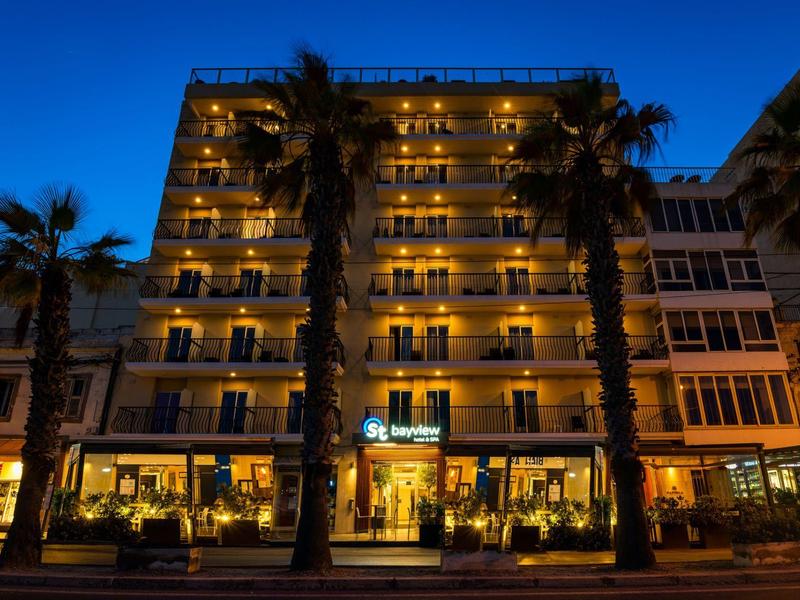 Mehrstöckiges Hotelgebäude mit beleuchteten Balkonen und Palmen vor blauem Abendhimmel.