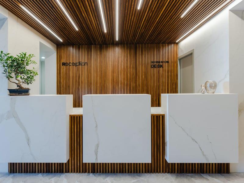 Moderne Rezeption mit weißer Marmoroberfläche, Holzrückwand und grüner Bonsai-Pflanze links.