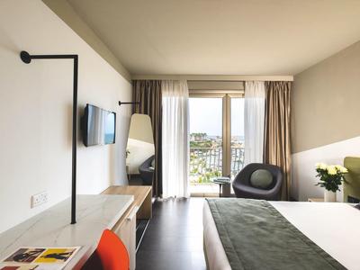 Camera d'hotel moderna con letto matrimoniale, scrivania, TV e balcone con vista sulla città.