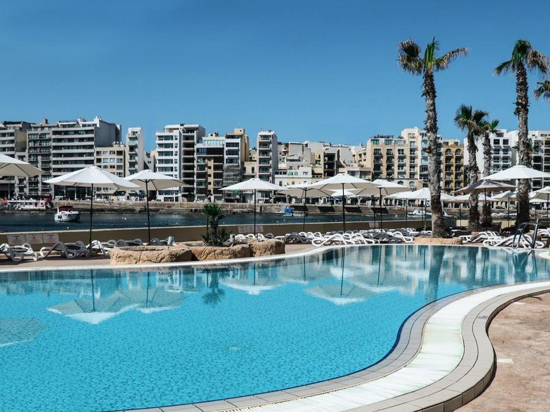 Grande piscina dell'hotel con ombrelloni e edifici sullo sfondo sotto un cielo sereno.