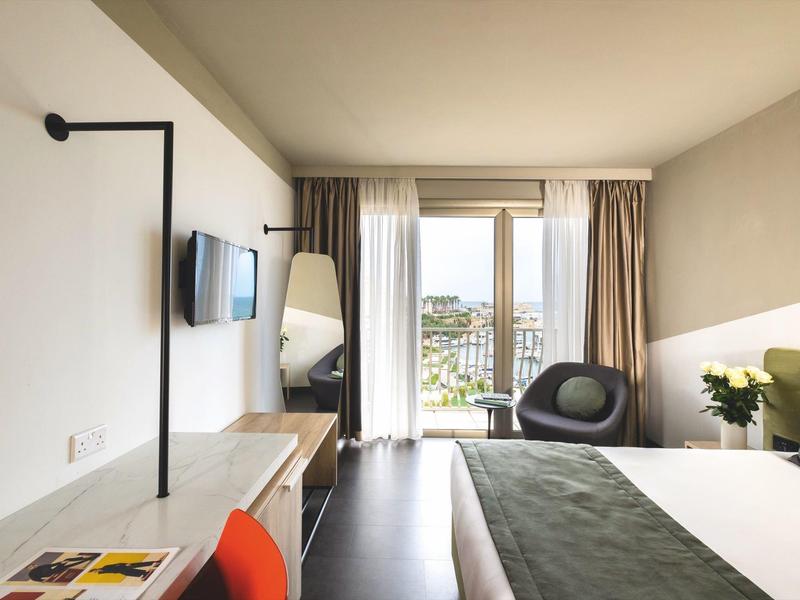 Camera d'hotel moderna con letto matrimoniale, scrivania, TV e balcone con vista sulla città.