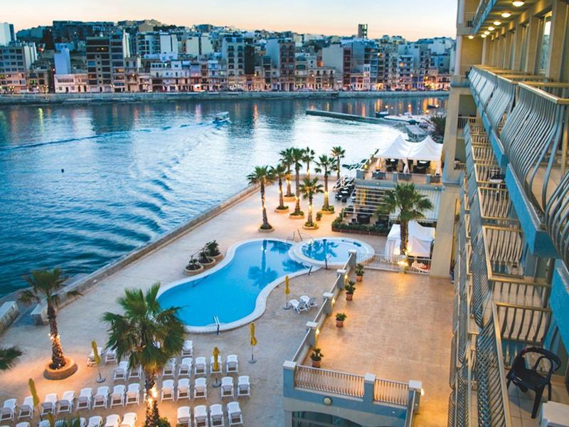 Vista di una piscina dell'hotel con palme e una promenade sul lungomare al tramonto.