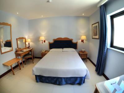 Lichte hotelkamer met tweepersoonsbed, bureau, stoel, nachtkastjes en raam met blauwe gordijnen.