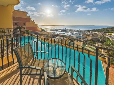 Balkon mit Stühlen und Tisch, Blick auf Hotelpool und Meer bei Sonnenuntergang.