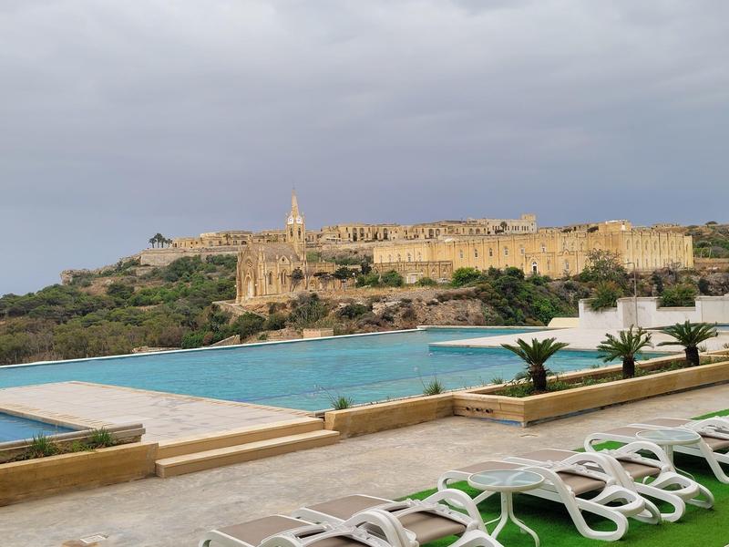 Zwembad met ligstoelen en uitzicht op een historische vestingstad bij bewolkte lucht.