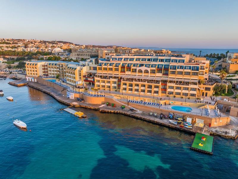 Hotelgebäude am Meer mit Bootsanleger, Wohnhäusern im Hintergrund und klarem blauem Wasser.
