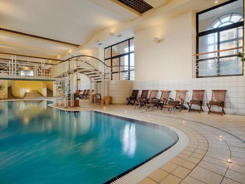 Innenraum mit großem Schwimmbecken, beigen Fliesen, Sitzstühlen und großem Fenster mit natürlichem Licht.