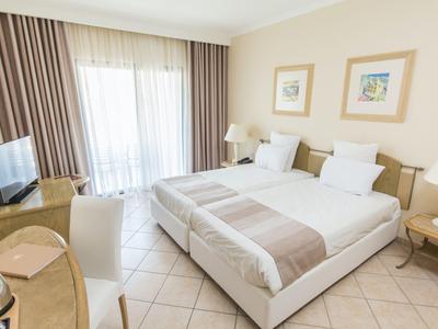 Helles Hotelzimmer mit zwei Einzelbetten, Schreibtisch, Fernseher und Balkon mit Vorhängen.