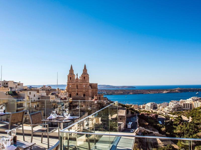 Coastal town with historic church, modern rooftop terrace, and clear blue skies over the sea.