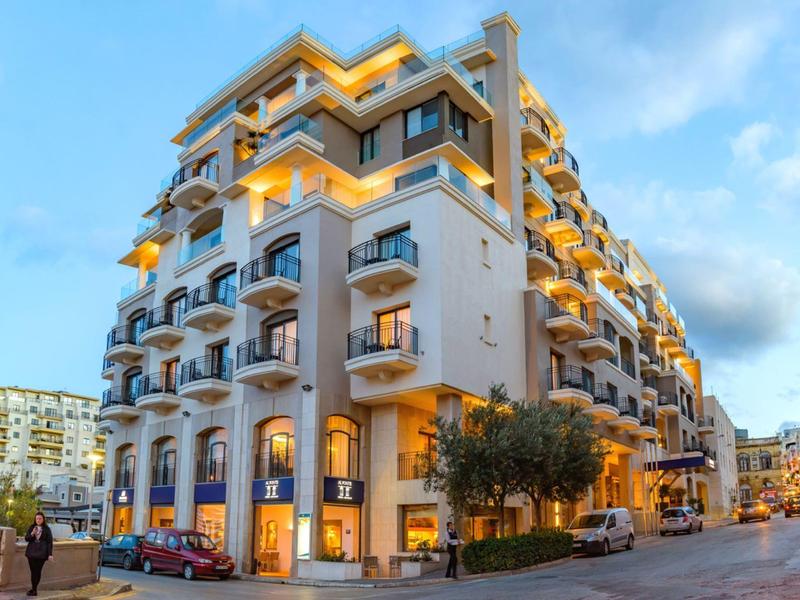 Modern hotel building with balconies and evening lights at street corner under blue sky.